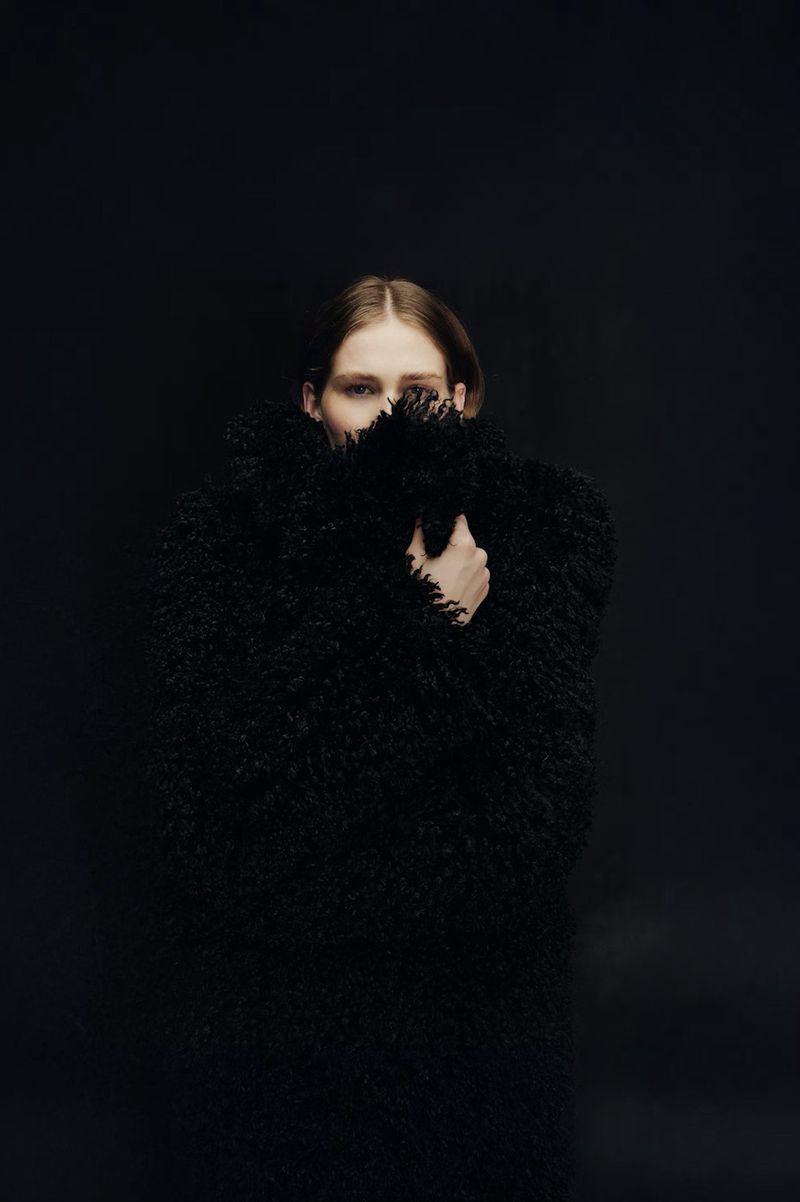 A fair-skinned woman with red hair poses in a black furry coat against a black background, with only her eyes visible.