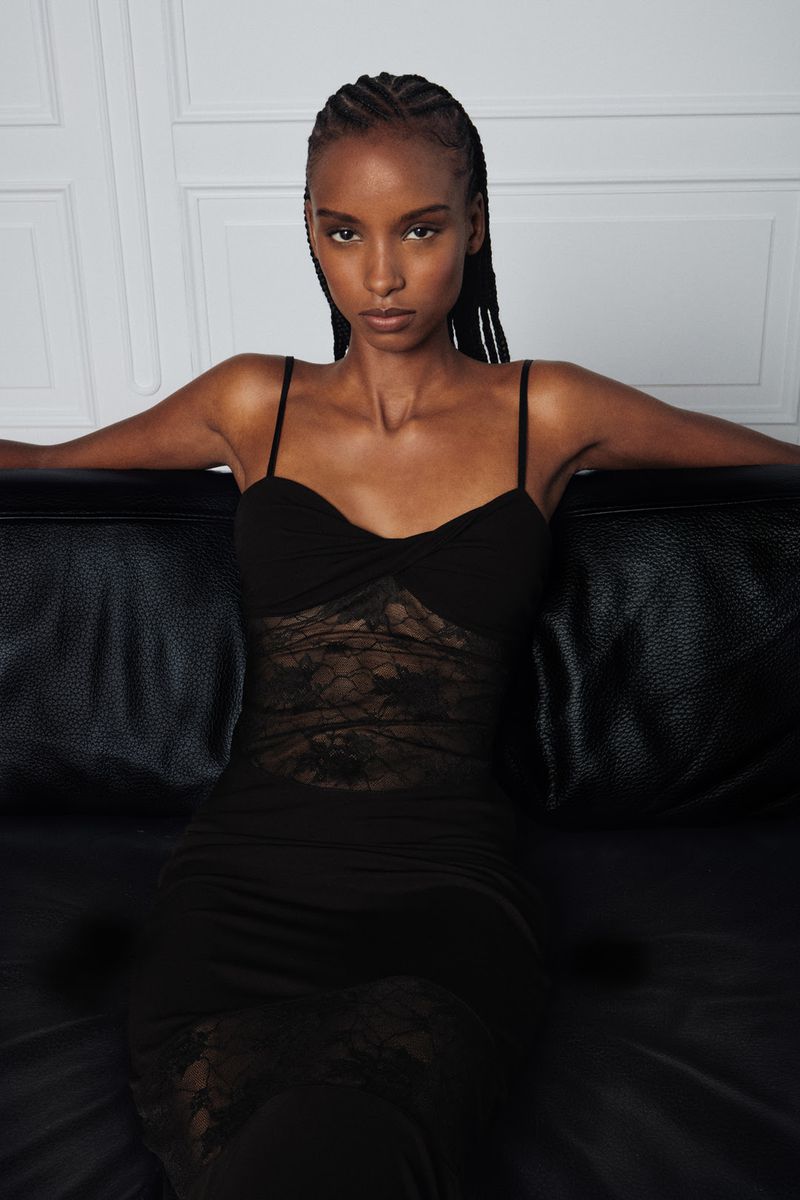 An African American woman poses in a black, semi-sheer dress on a black leather couch against a white wall, showcasing fashion and style.