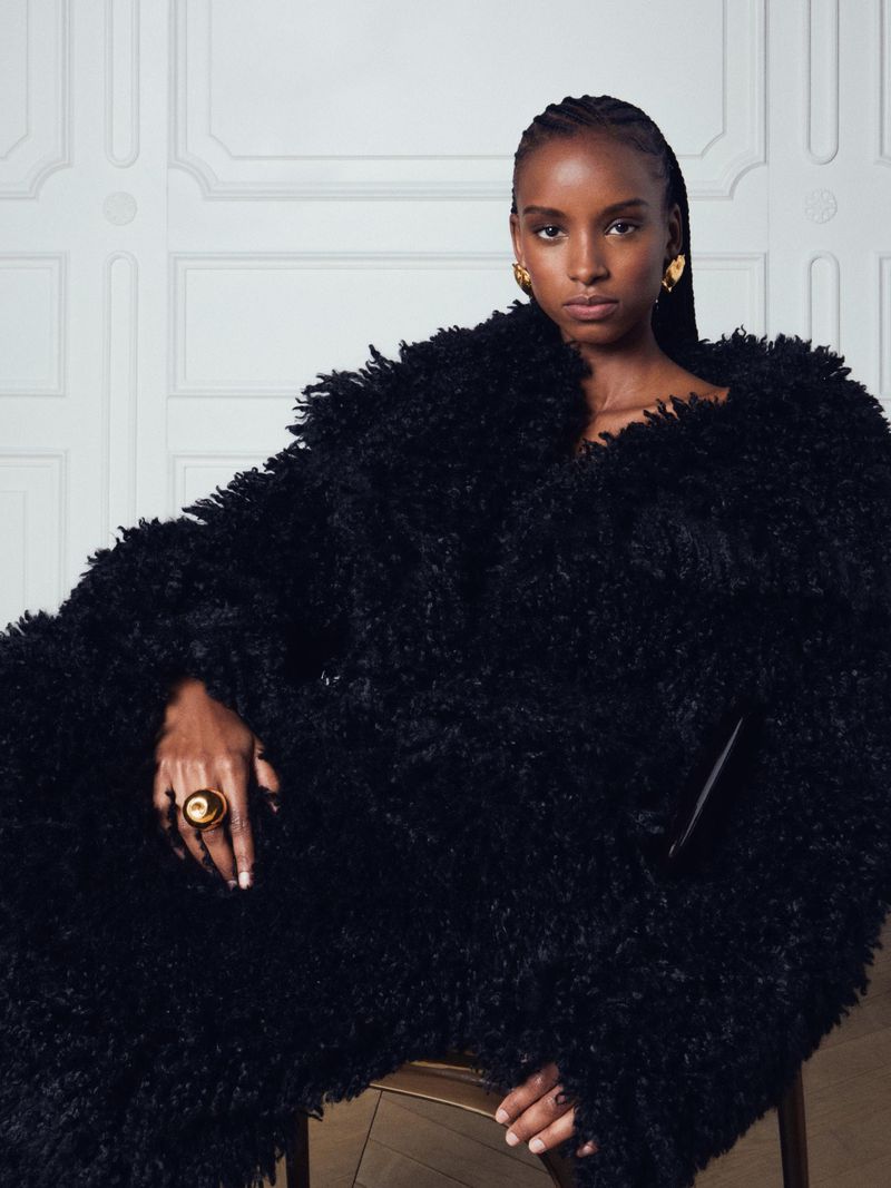 Model wearing a black textured coat seated against a white paneled wall for Nina Ricci SS25