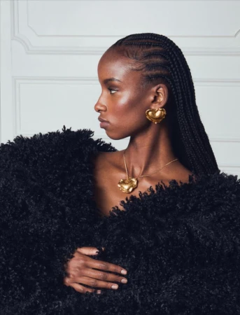 Profile shot of a dark-skinned woman with braids, wearing gold earrings and a necklace, with a dark furry coat against a white wall.