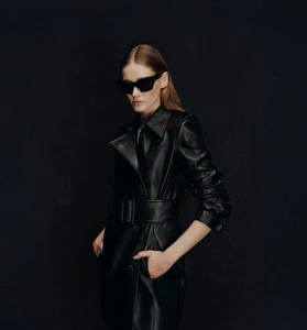 Model wearing dark sunglasses and a black leather belted coat against a dark textured studio backdrop for Nina Ricci SS25.