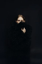 Model in textured black fuzzy coat against a dark seamless backdrop with hands partially covering the face