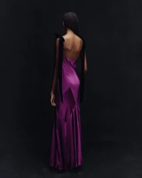 Model from behind wearing a glossy purple silk slip gown with black shoulder ribbons against a dark seamless studio backdrop.