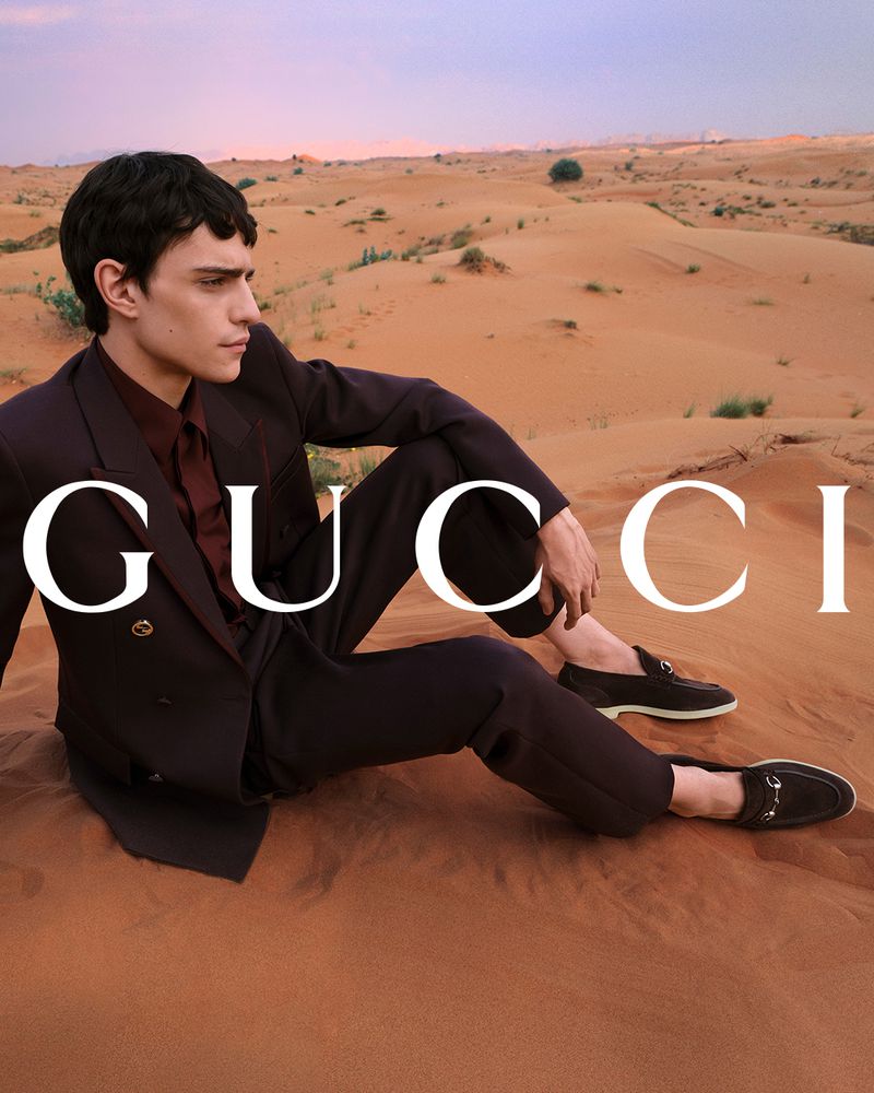 Man poses in Gucci suit and loafers, sitting in a desert landscape with sand dunes, under a soft sunset sky with the Gucci logo.