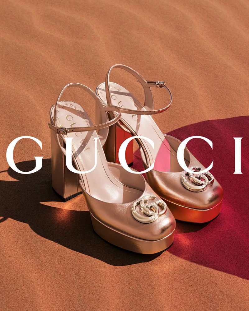 Elevated view of pair of gold Gucci platform heels with ankle straps, accented by red and tan ground, and partial brand name.