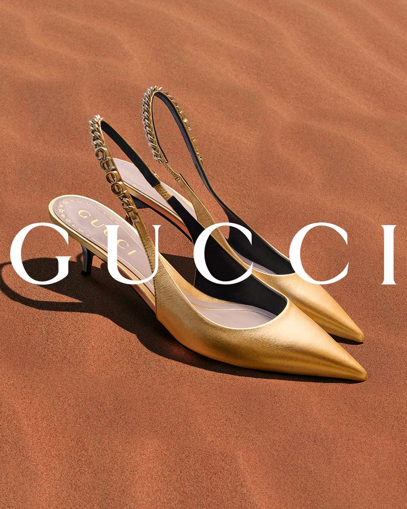 Two gold-colored Gucci slingback high heels with chain detail displayed against a brown background in a fashion advertisement.