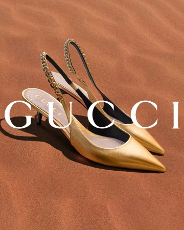 Two gold-colored Gucci slingback high heels with chain detail displayed against a brown background in a fashion advertisement.