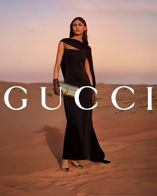 Fashion shot of a woman in a black dress and gold shoes, holding a gold purse, against a desert backdrop with a large brand logo.