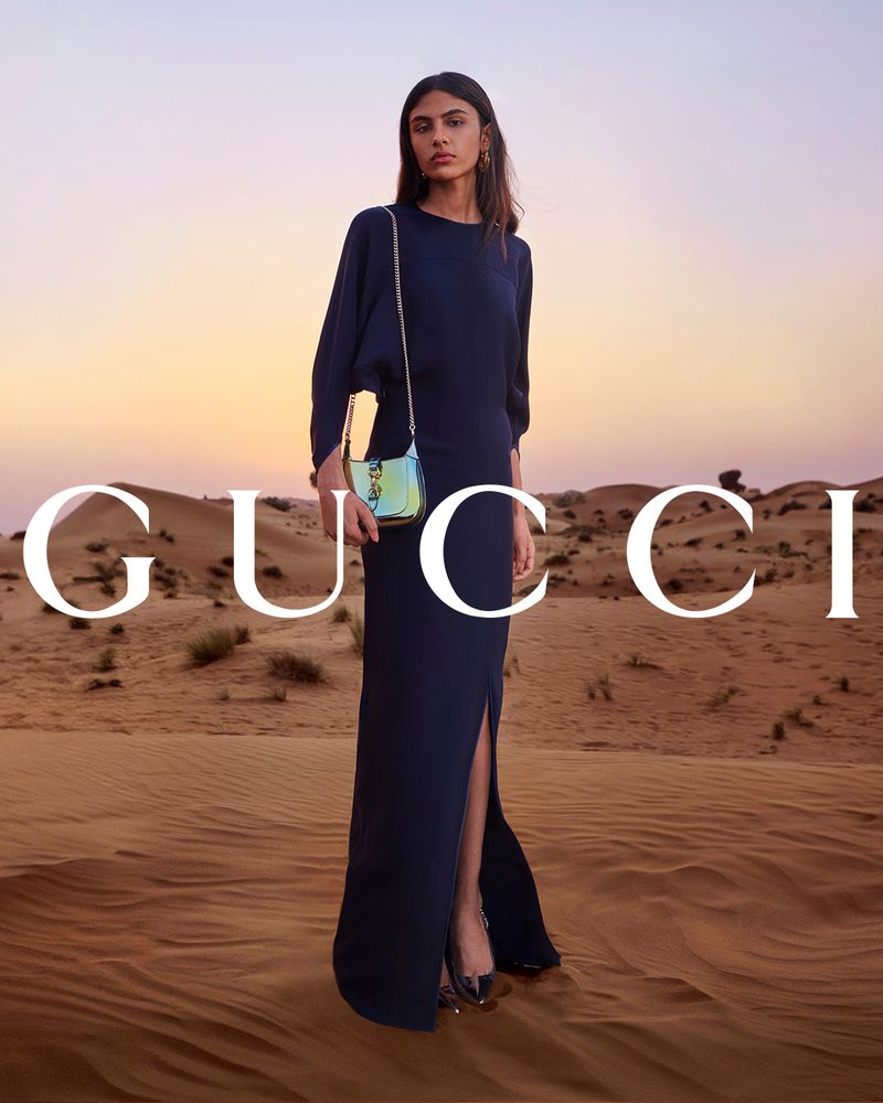 Elegant dark blue gown showcased in a desert landscape at sunset, paired with a metallic shoulder bag, part of a fashion editorial.