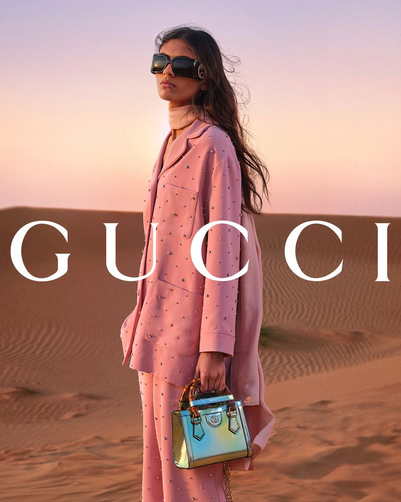Fashion shot of a woman in a pink suit holding a Gucci handbag standing in a desert during golden hour with the Gucci logo visible.