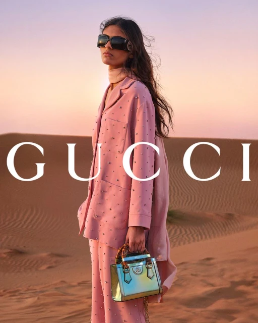 Fashion shot of a woman in a pink suit holding a Gucci handbag standing in a desert during golden hour with the Gucci logo visible.