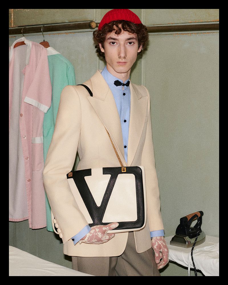 A male model wearing a beige suit, red knit hat, and lace gloves poses with a Valentino bag on a white ironing board.