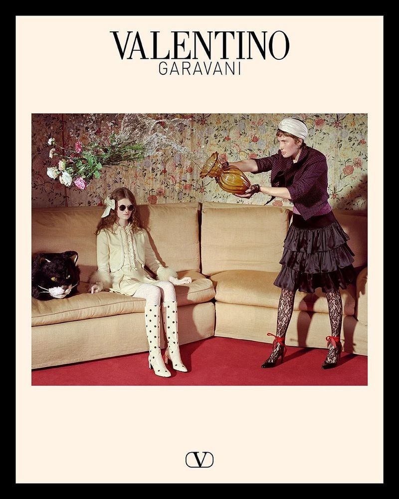 Two fashion models pose indoors for Valentino, one sitting on a beige couch, the other standing holding a vase, floral wallpaper backdrop.