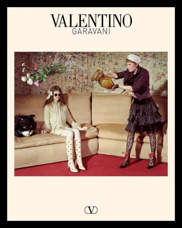 Two fashion models pose indoors for Valentino, one sitting on a beige couch, the other standing holding a vase, floral wallpaper backdrop.