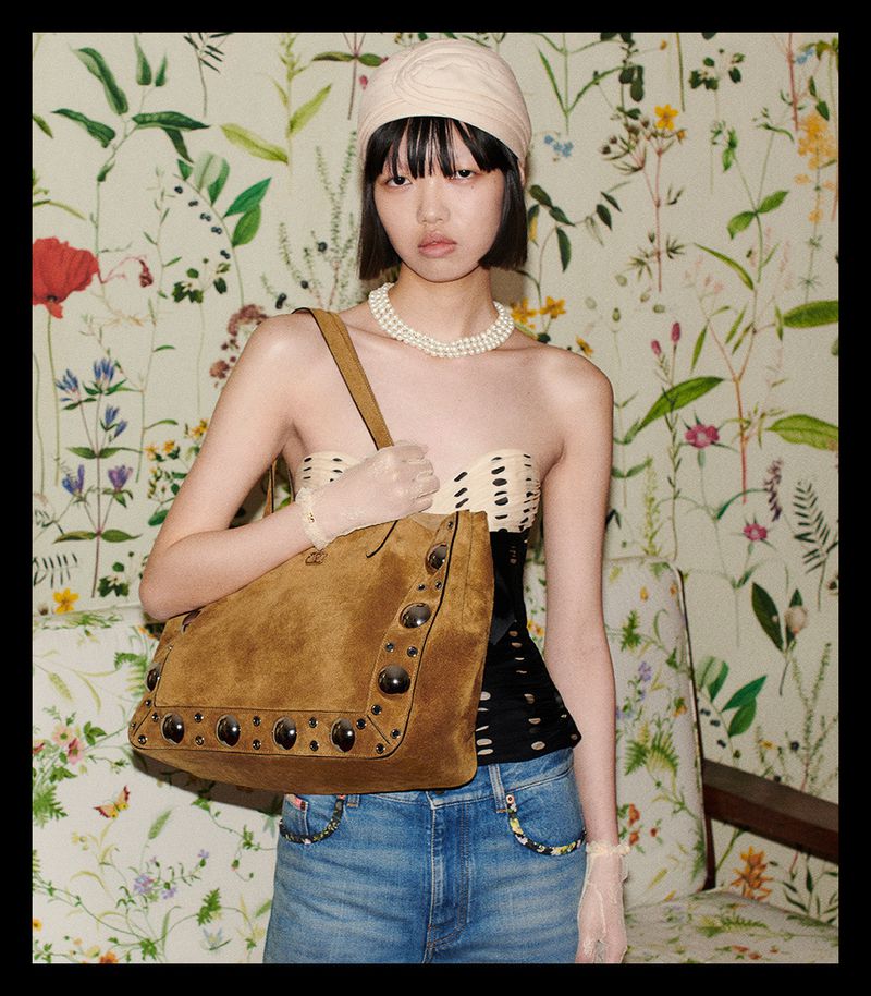 Fashion shot of a young woman with a suede bag, wearing a cream-colored top, light-wash jeans, a pearl necklace, and a turban, with a floral background.