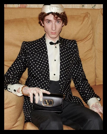 A slender man in a polka dot blazer, black bow tie, and cream-colored headwrap sits on a sofa holding a small black purse.
