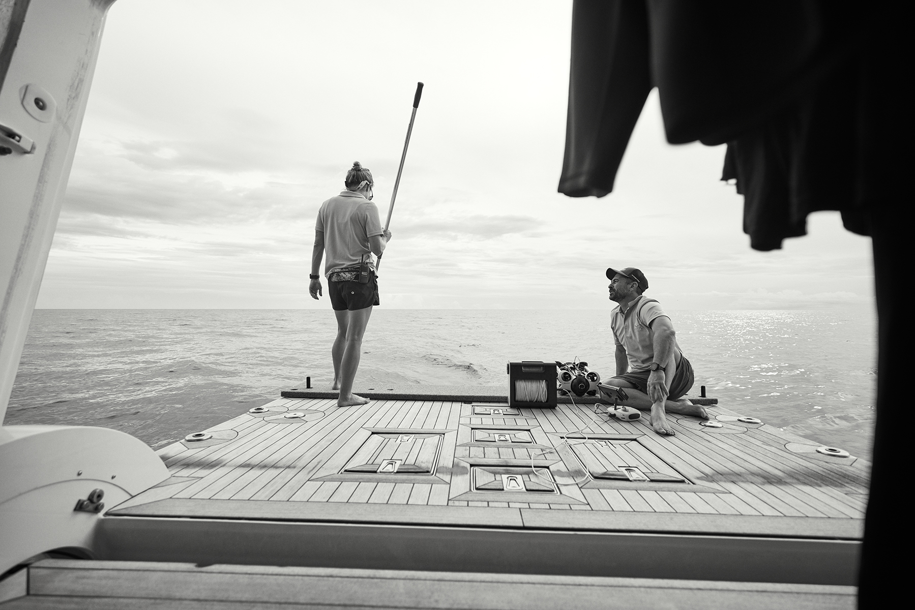 Two people on the back of a boat on the ocean with a cloudy sky. One person is standing and the other is sitting near equipment.