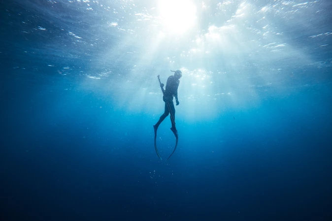 Underwater photo of a free diver swimming in clear blue ocean water with sunlight shining from above creating a serene and peaceful scene.