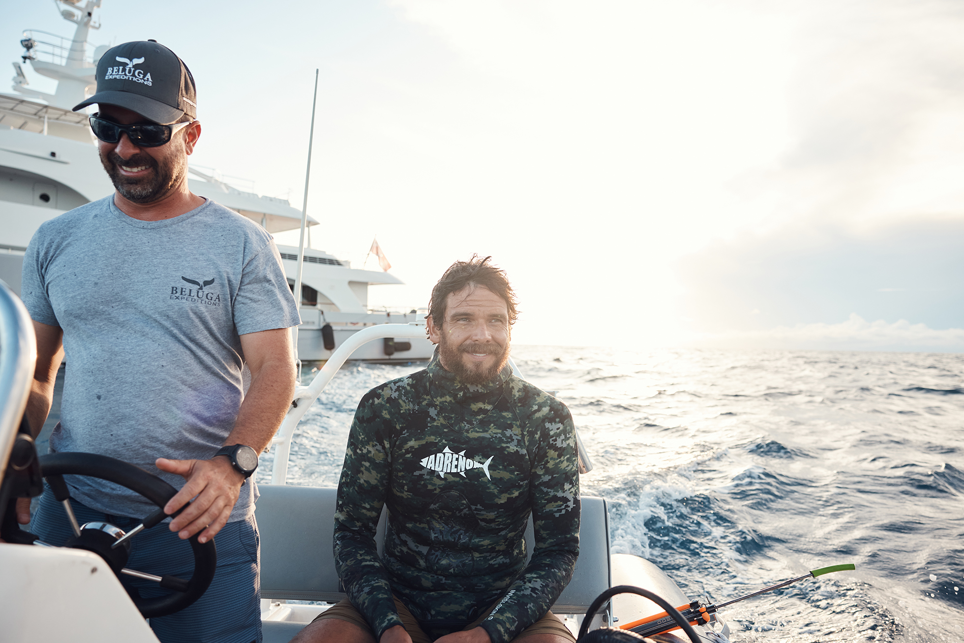 Two men aboard a motorboat on the ocean with a luxury yacht in the background, enjoying a sunny day on the water.