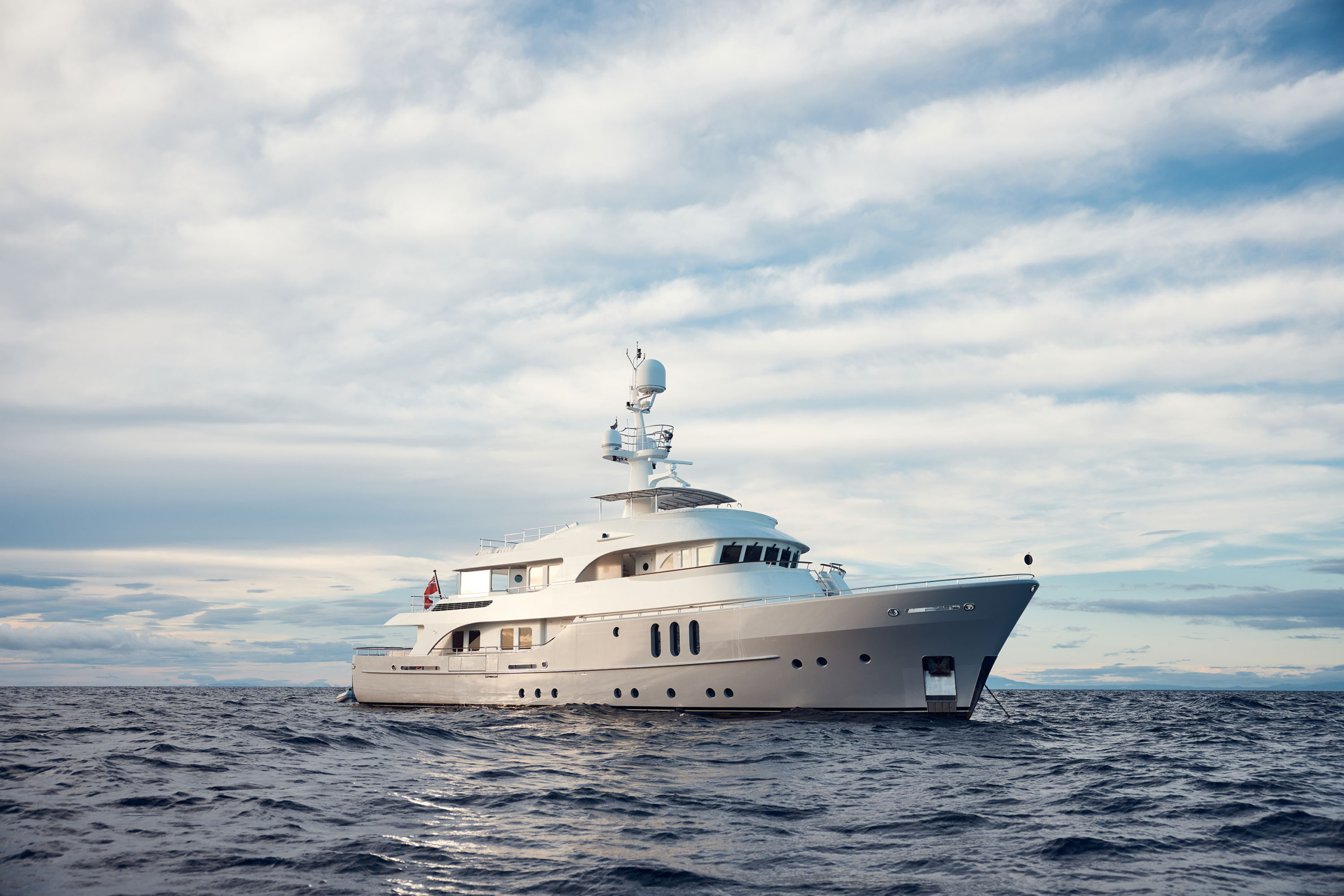 Luxury yacht on calm ocean waters against a cloudy sky. The yacht is predominantly white with visible deck and antenna details.