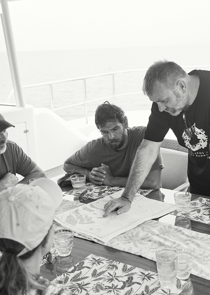 Four people gathered around a table on a boat, reviewing a map; monochrome image. One person pointing at the map. Water visible.