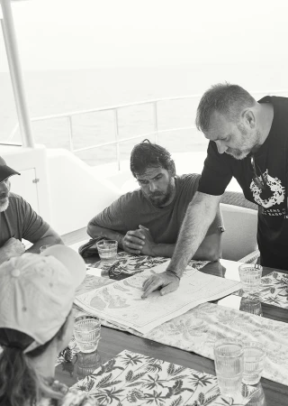 Four people gathered around a table on a boat, reviewing a map; monochrome image. One person pointing at the map. Water visible.