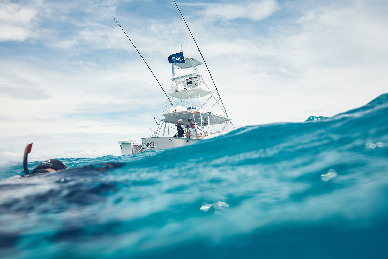 Fishing boat with anglers on open ocean beneath cloudy sky, scuba diver partially submerged in turquoise water in foreground.