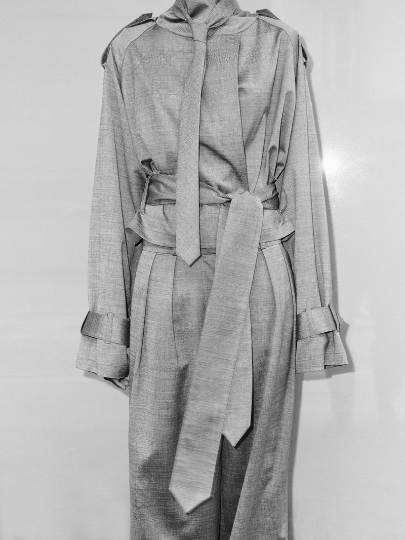 Monochrome image of a light grey, fabric trench coat with tie belt and long sleeves, presented against a plain, light grey backdrop.
