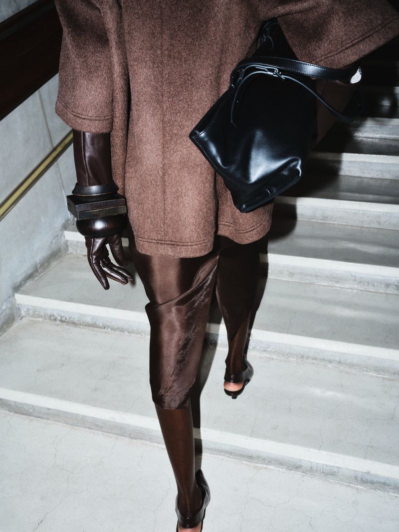 Fashionable woman walks down stairs wearing brown coat dress, high-fashion black purse, oversized gloves, and brown thigh-high boots.