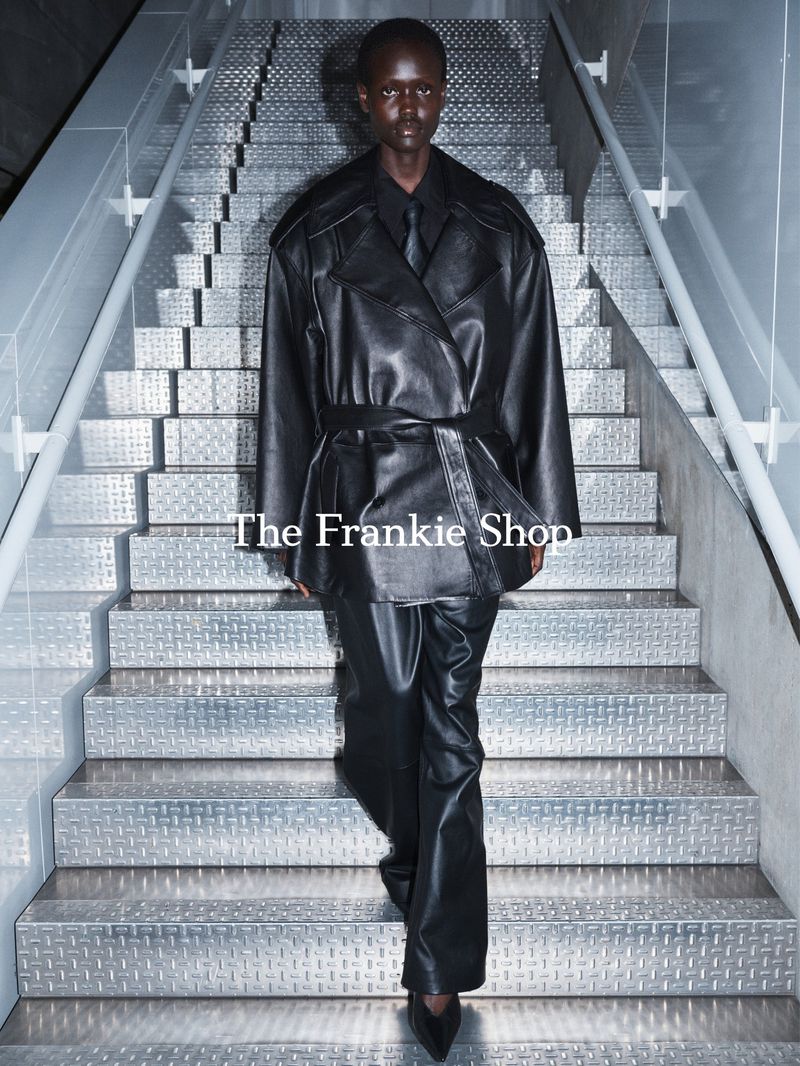 Fashionable woman in a belted leather trench coat and matching pants walks down gray metal stairs in a modern indoor setting.