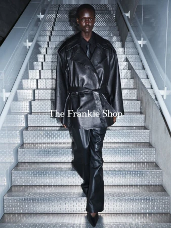 Fashionable woman in a belted leather trench coat and matching pants walks down gray metal stairs in a modern indoor setting.