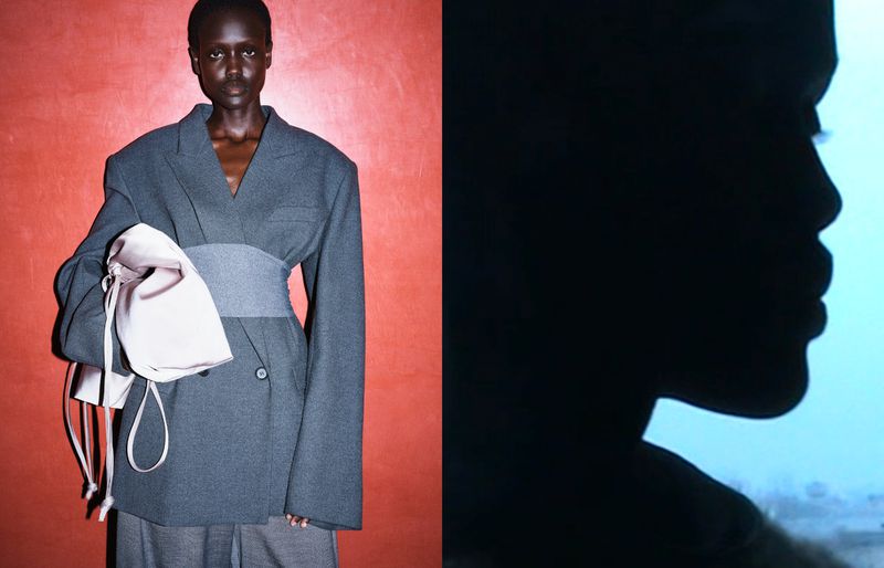 Dark-skinned female model in an oversized grey blazer, cinched waist, holding a satin bag, against a red backdrop; profile silhouette right.