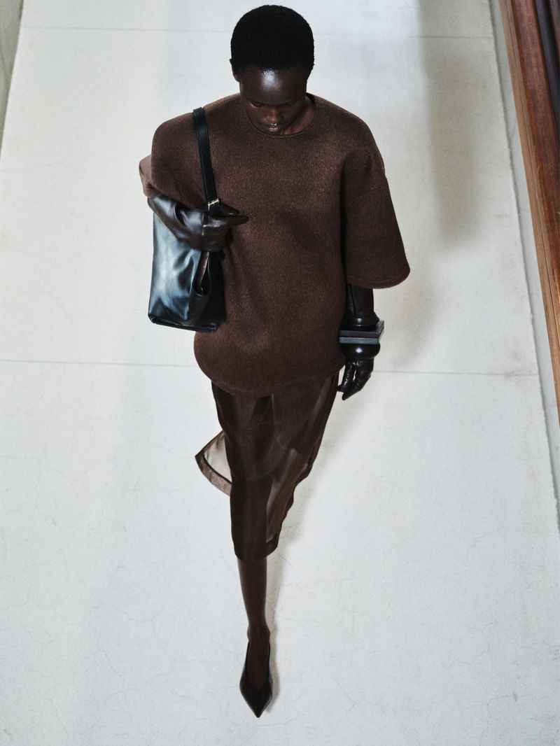Runway fashion: a Black model presents a brown dress with a black tote bag in a minimalist interior setting, captured from a high-angle perspective.