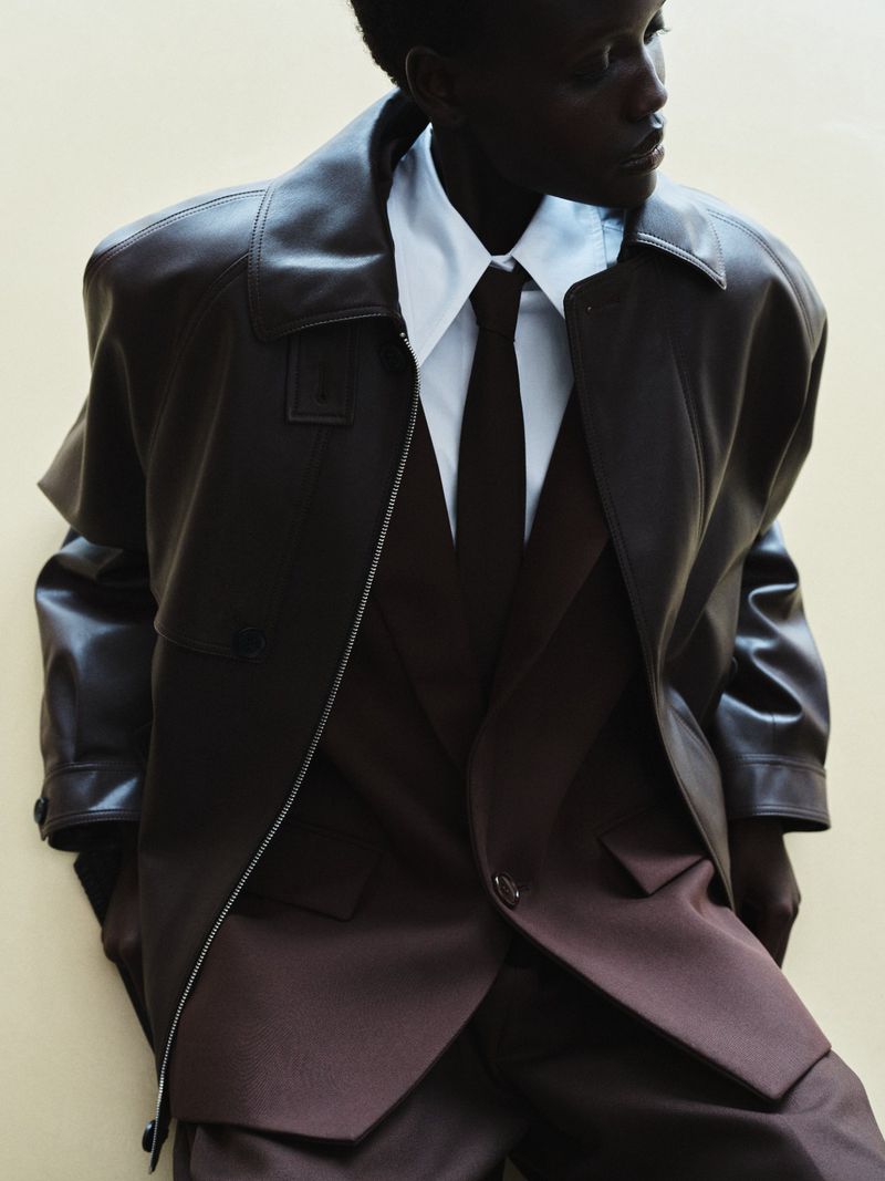 Dark-skinned adult posing indoors in a brown suit, white collared shirt, and brown leather coat. Neutral wall background.
