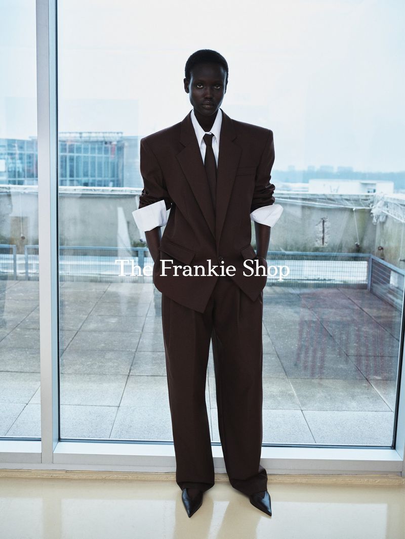 A slender Black model stands near a window wearing a brown suit with wide shoulders, white cuffs, and black heeled shoes in a studio.