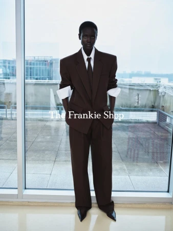 A slender Black model stands near a window wearing a brown suit with wide shoulders, white cuffs, and black heeled shoes in a studio.