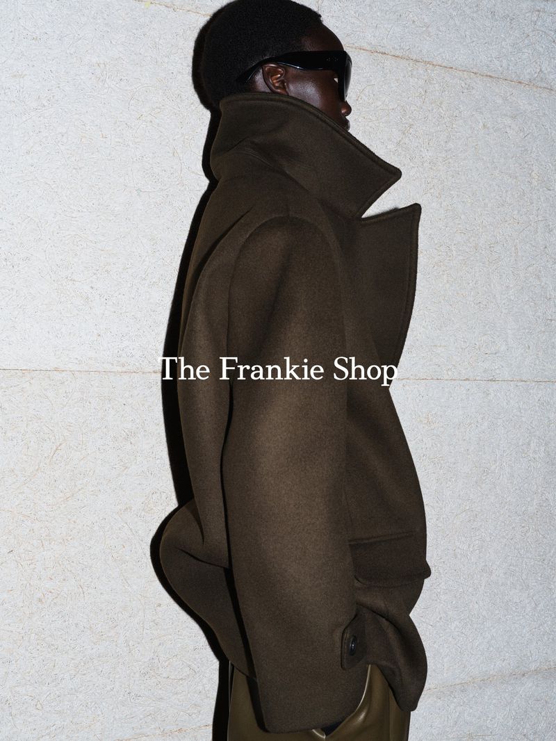 Model in dark sunglasses and olive green high-collar coat against a textured pale wall.