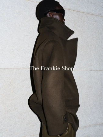 Model in dark sunglasses and olive green high-collar coat against a textured pale wall.