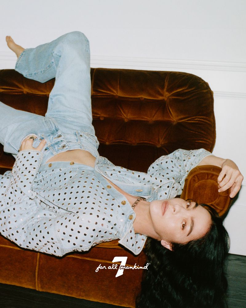 Model lying on a brown velvet tufted sofa wearing a light wash denim set against a plain white wall