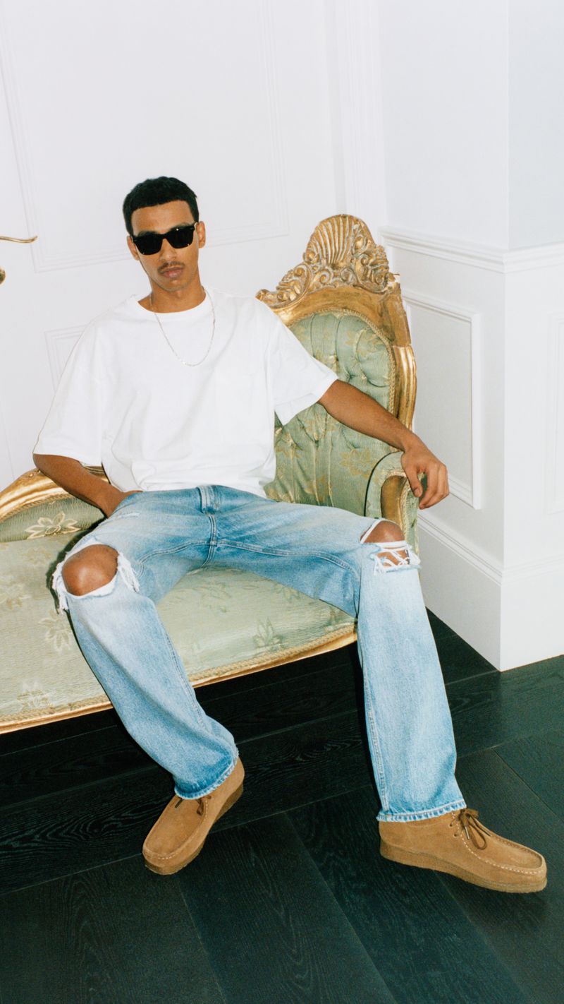 Man in a white t-shirt, distressed denim jeans, and brown suede loafers poses on a gold and green settee with white walls.