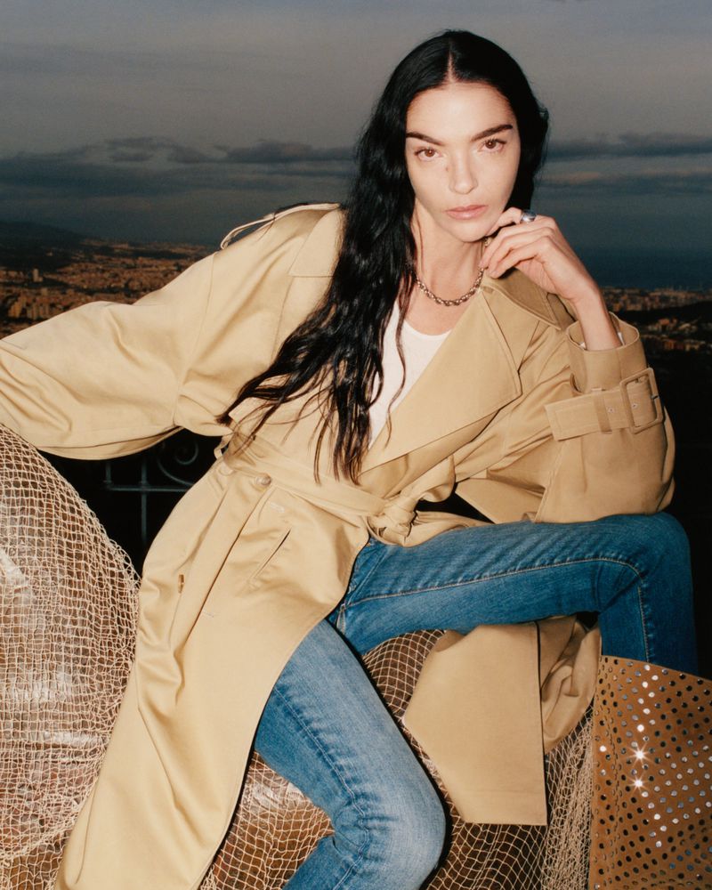 Mariacarla Boscono wearing a beige trench coat and blue jeans against an outdoor landscape background for 7 For All Mankind
