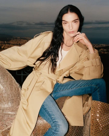 Mariacarla Boscono wearing a beige trench coat and blue jeans against an outdoor landscape background for 7 For All Mankind