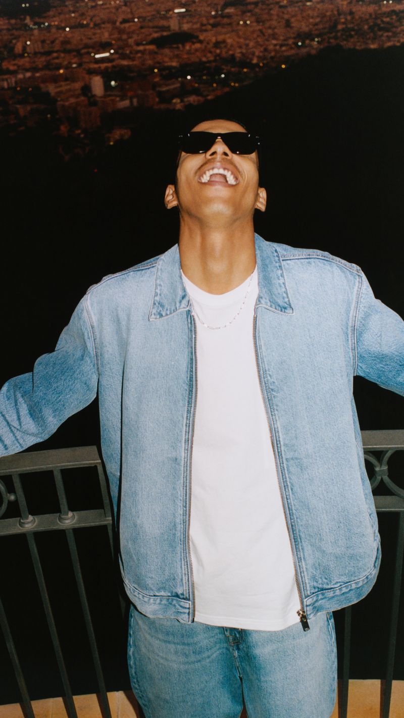Young man in denim jacket with arms outstretched, wearing sunglasses, stands in front of illuminated cityscape at night.
