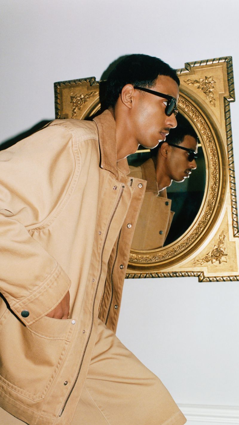 Stylish man in a tan outfit and sunglasses poses indoors, with a mirror reflecting his image in a fashion editorial shot.