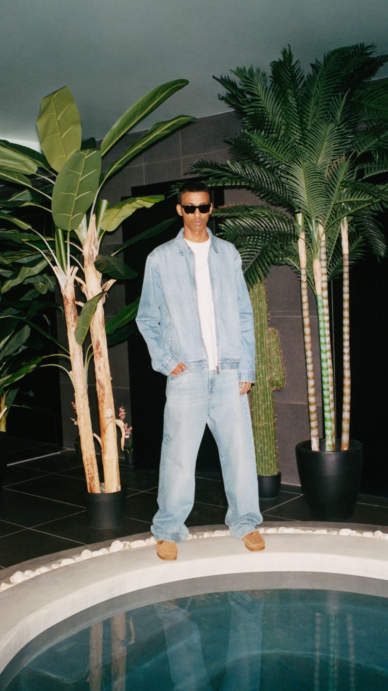 Man in a light denim jacket and pants standing in front of an indoor pool, framed by two large potted tropical plants.