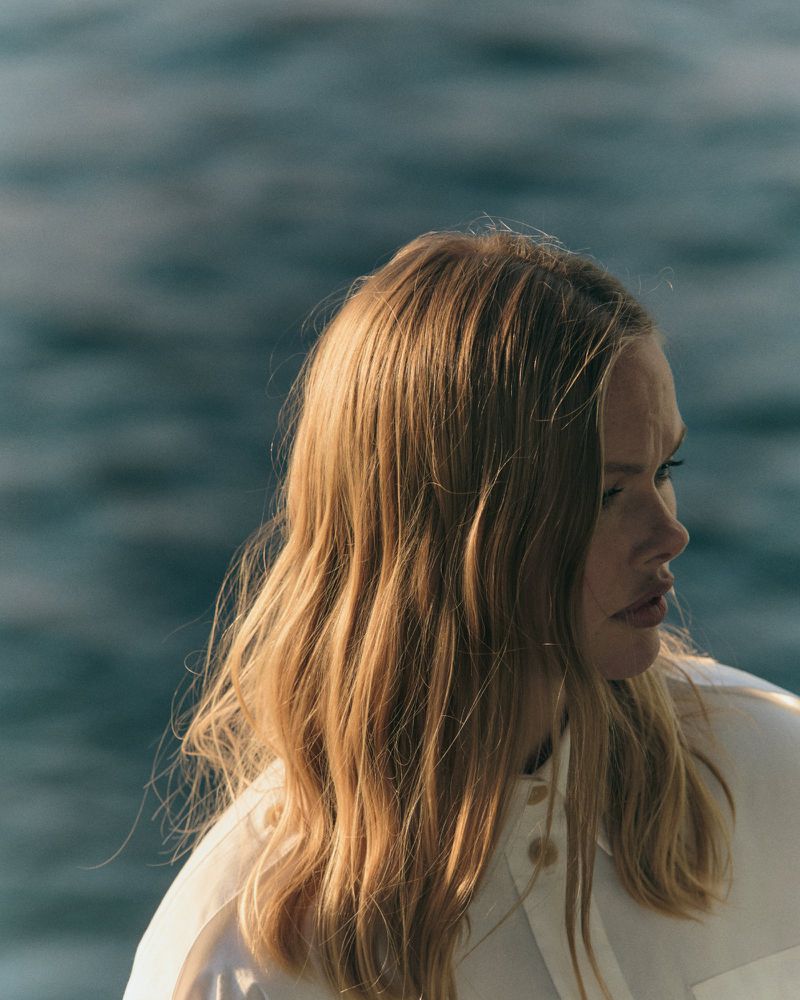 Close-up profile of a woman with blonde hair wearing a white collared shirt against a rippling water background