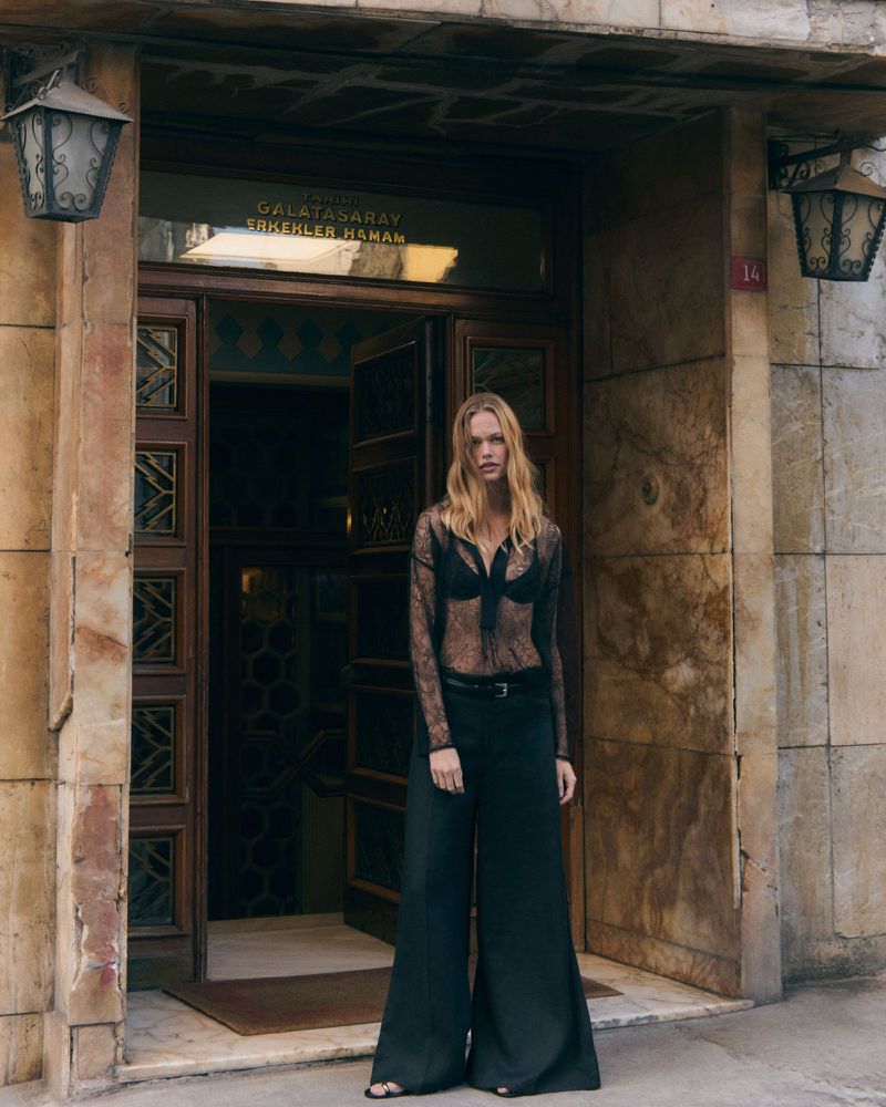 Model standing in front of an ornate wooden doorway wearing a sheer black lace top and wide-leg trousers