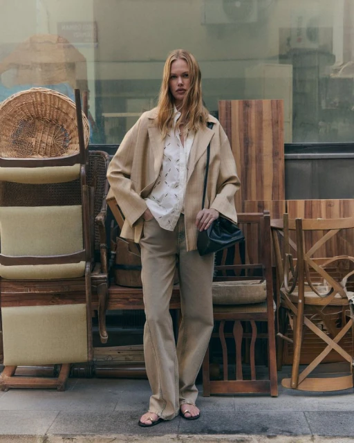 Fashionable adult female poses outdoors in a beige jacket, patterned top, and flared jeans, holding a black purse near stacked wooden chairs.