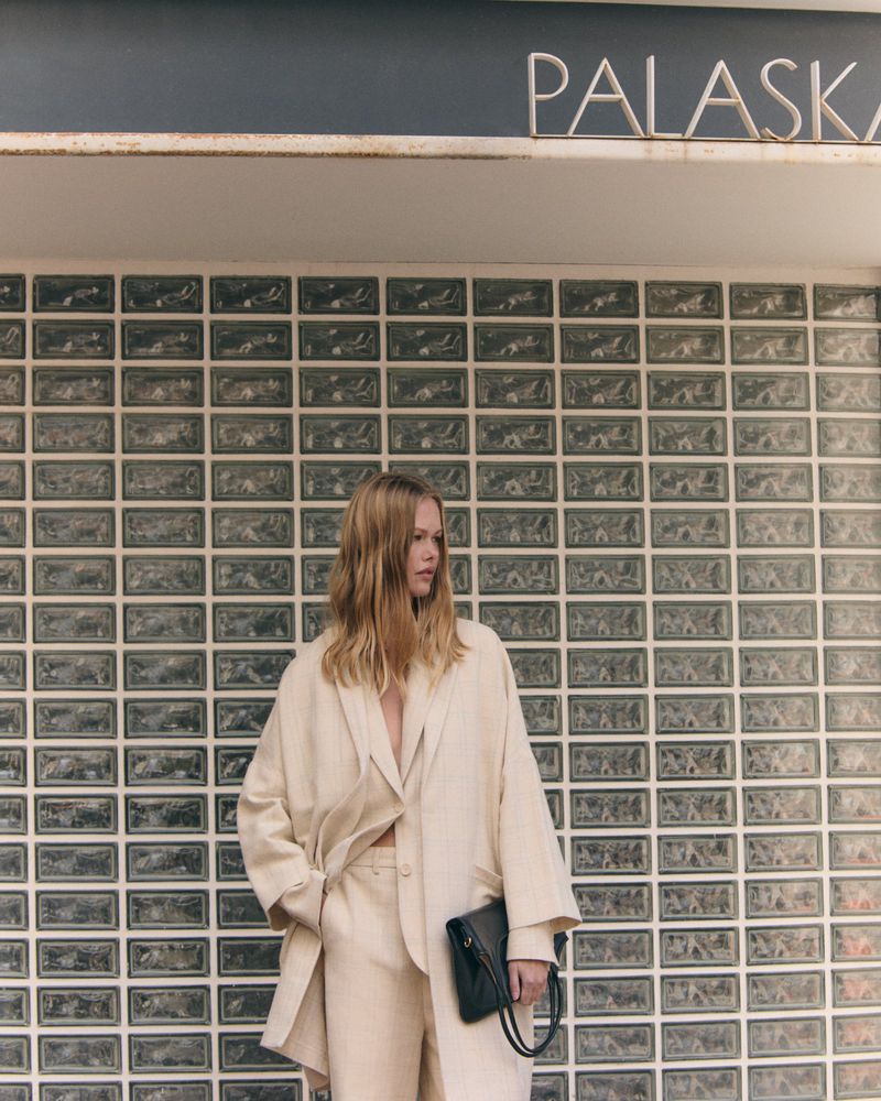 Woman in a cream-colored suit stands in front of a textured wall, holding a black handbag. The setting includes a sign above.