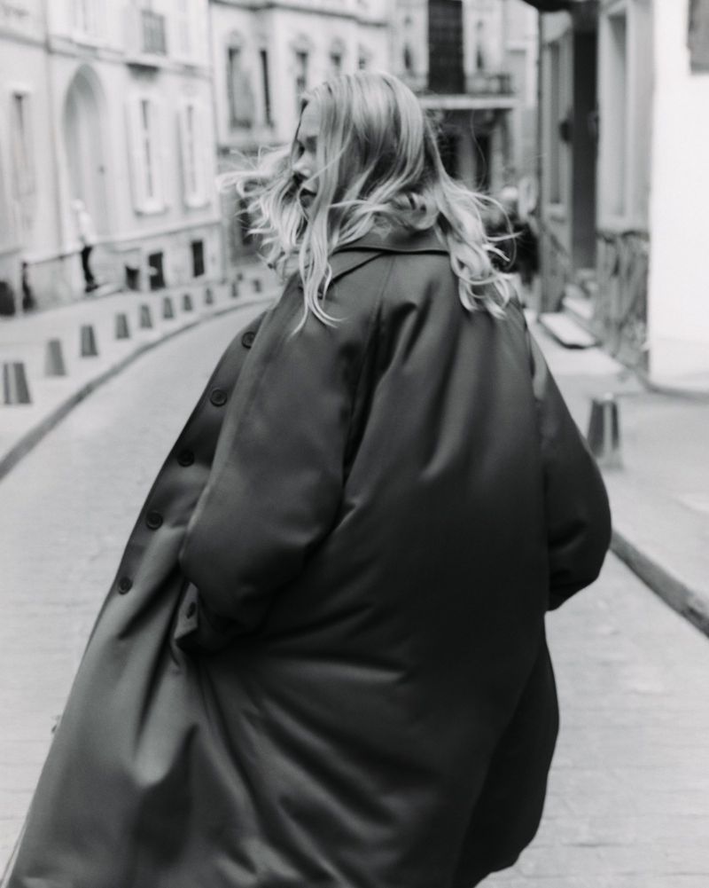 Black and white shot of a woman with long hair walking on a city street wearing an oversized coat in an urban setting.
